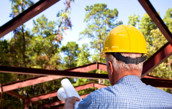 Man in a yellow hard hat watches as a steel building frame is lowered into place