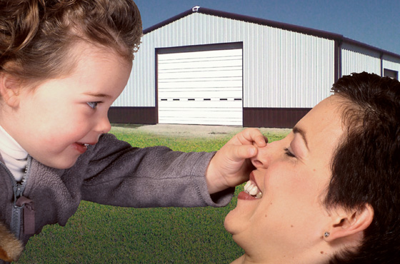 Mother plays with toddler before a white steel building with brown trim