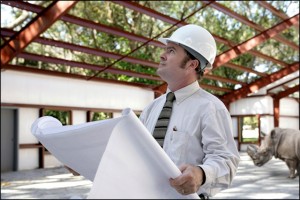 A man in a tie and hard hat holds building plans as he gazes upward through the steel framing of a building under construction